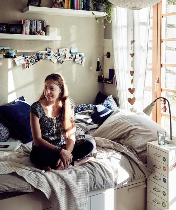 Malin assise sur son lit recouvert d’un linge de lit aux tons neutres et de coussins bleus et au-dessus duquel se trouvent des étagères suspendues au mur.