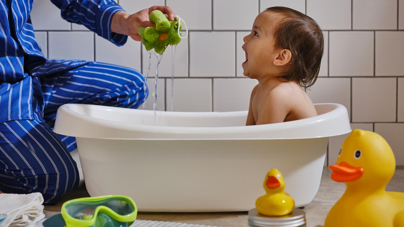 Malo dete u beloj LÄTTSAM bebi-kadici, sa zidom s belim pločicama iza, smeje se dok odrasla osoba drži zeleni sunđer.