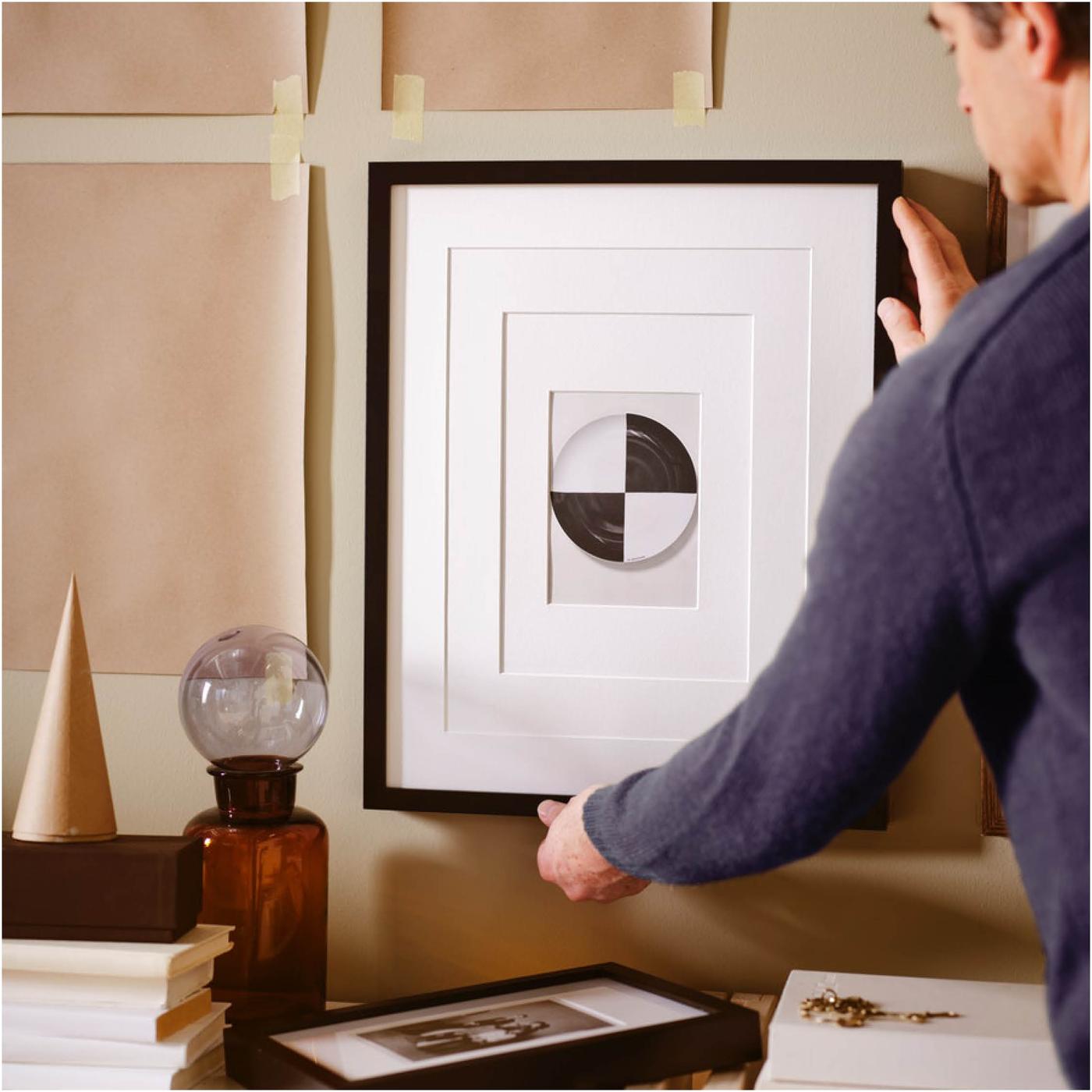 Man hangt een afbeelding in een zwarte RIBBA lijst aan een beige muur waarop bruine papieren mallen getapet zijn.