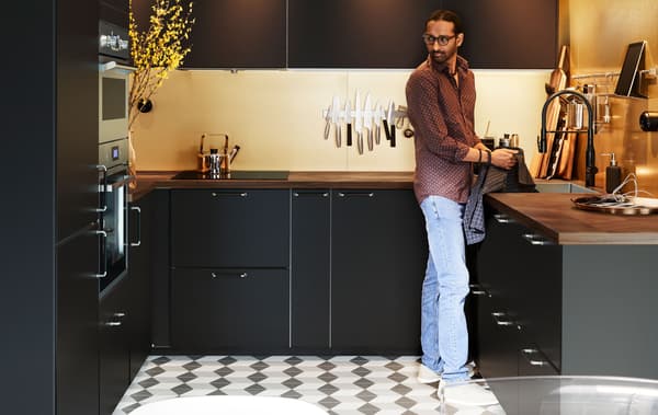 Man standing in a clean, modern U-shaped kitchen with dark cabinets, checkered floor tiles, and subtle lighting.