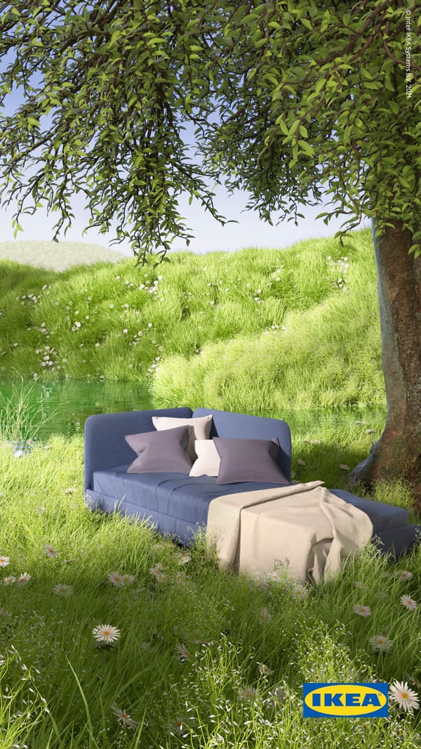 Medium-blue BLÅKULLEN bed frame with corner headboard, a throw and cushions on top, stands by a pond among lush green grass.