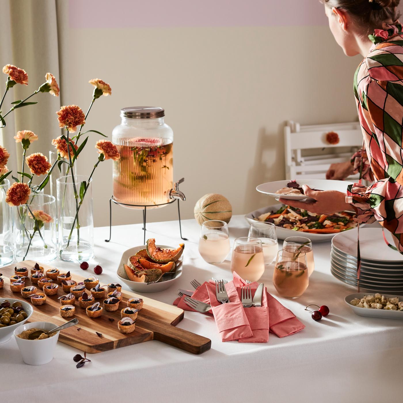 Mesa de bufê com vários pratos e um recipiente com torneira VARDAGEN com uma bebida cor de pêssego a ser vertida em copos.