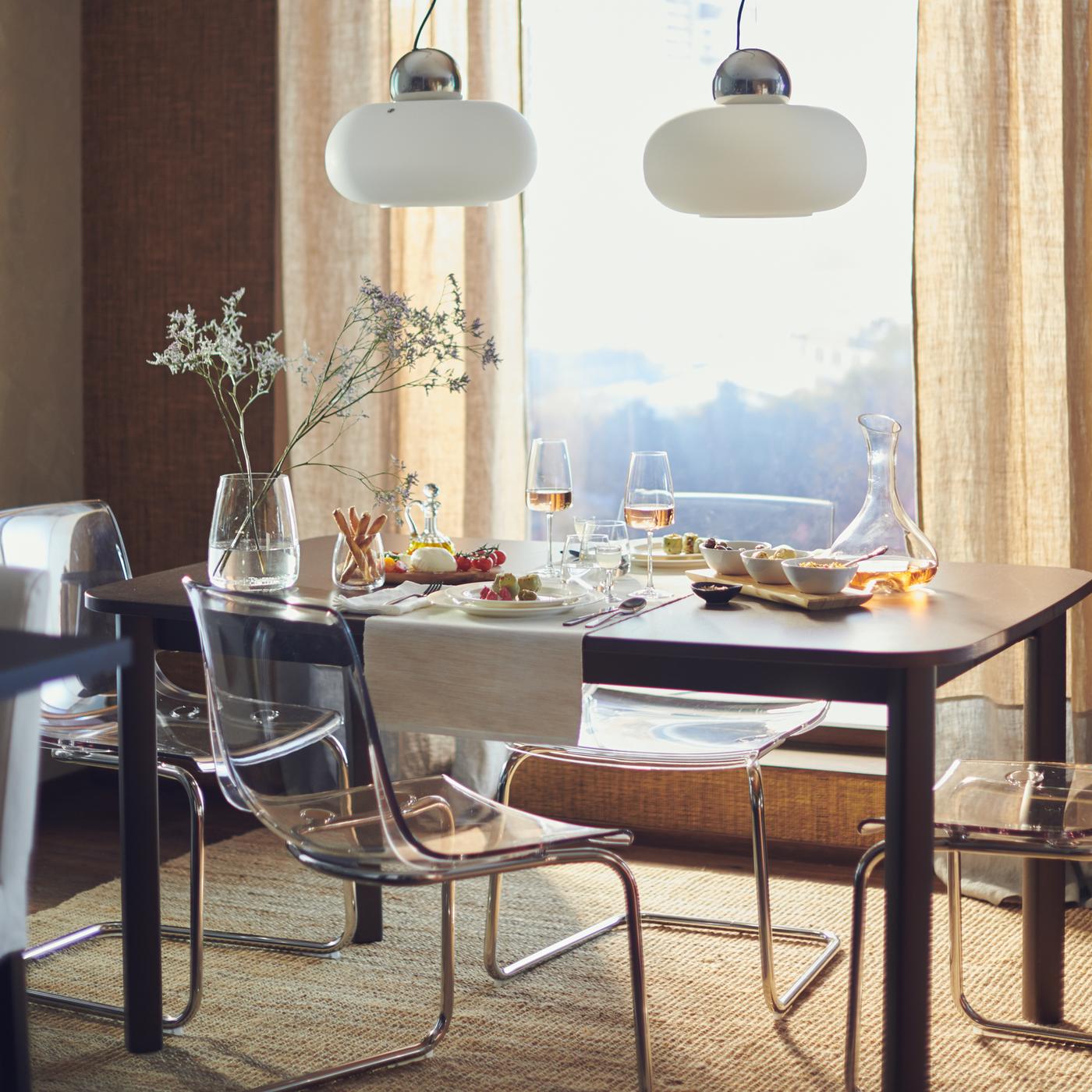 Mesa extensível STRANDTORP junto a uma janela grande e luminosa, posta para duas pessoas com comida, copos e uma jarra de flores.