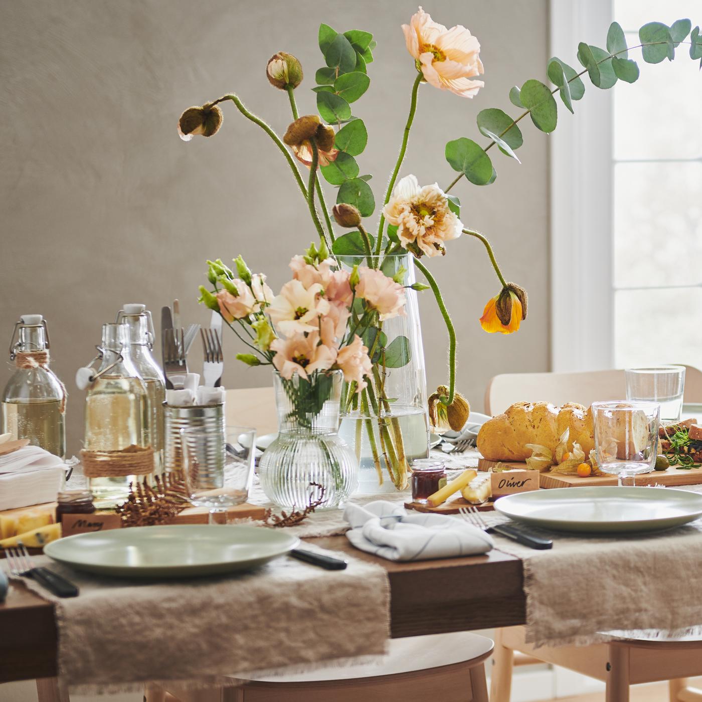 Mesa MÖRBYLÅNGA decorada para festa com tiras de tecido AINA, pratos FÄRGKLAR, flores em jarras e várias decorações.