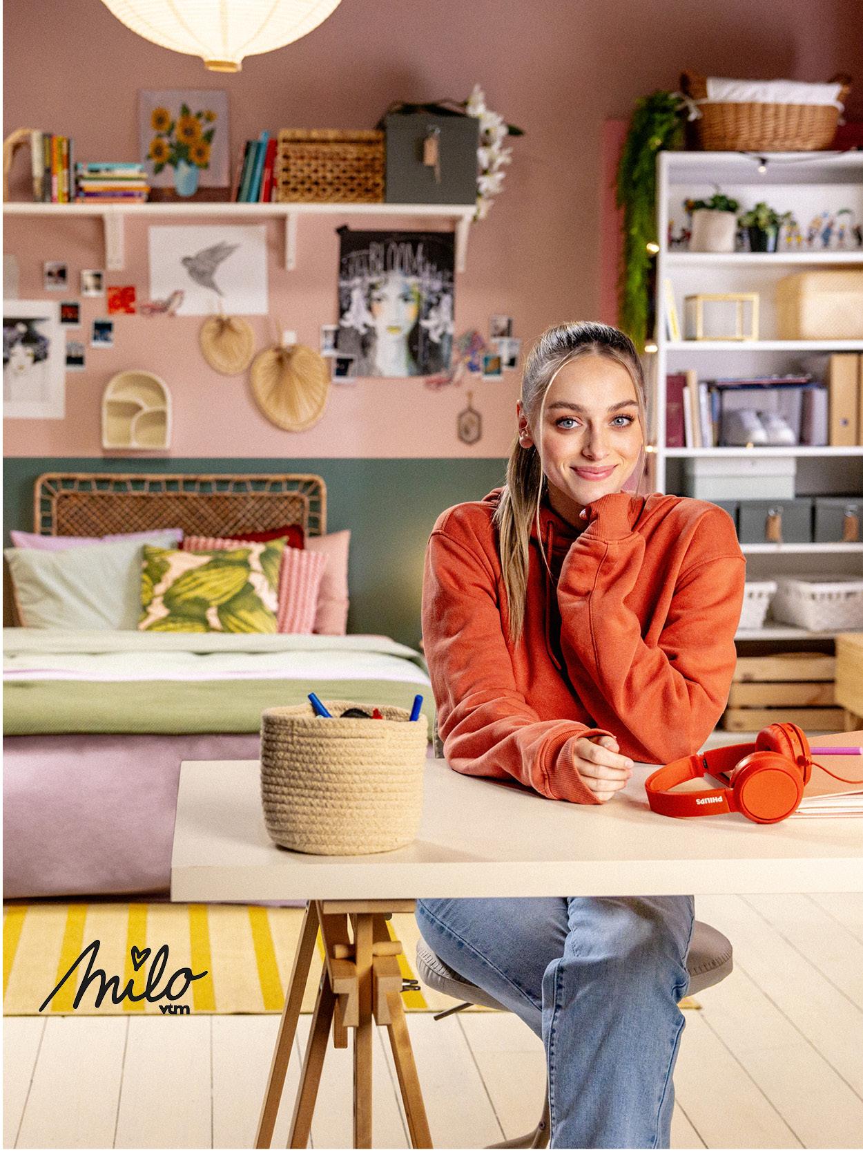Milo zit aan een IKEA tafel in een kleurrijke kamer met planten en decoraties, met een koptelefoon.