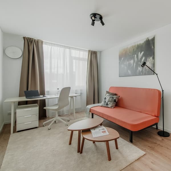 Minimalist home office with a bright orange sofa, geometric-patterned cushion, and black floor lamp. A modern desk setup with a white swivel chair, laptop, and drawer unit sits by a large window with beige curtains. Two oval nesting coffee tables and a soft beige rug add warmth to the space. A serene nature-themed canvas decorates the wall above the sofa.