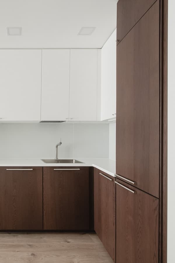 Minimalist kitchen corner featuring white upper cabinets and dark wood lower cabinets with sleek horizontal handles. A modern single-handle faucet is mounted on a white countertop above a stainless steel sink, with a light wood floor enhancing the clean, contemporary design.