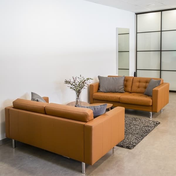 Minimalist office waiting area with two brown leather sofas, small wooden coffee table, and grey cushions on a dark rug.