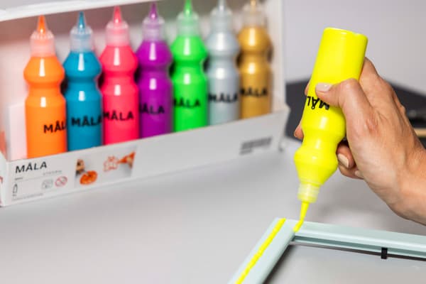 Mit MÅLA Farbe werden gelbe Linien auf einem Bilderrahmen gezeichnet.
