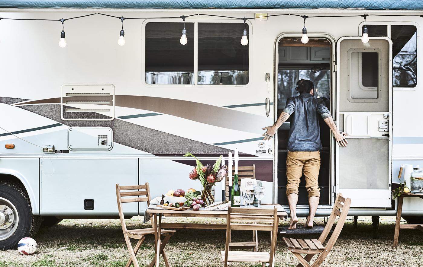 Mobilier d’extérieur installé devant un mobil-home décoré d’une guirlande lumineuse, avec Karlton debout devant l’entrée.
