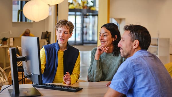 モダンなオフィスで、テーブルに座ってコンピューター画面を見ながら協力する3人の人物。