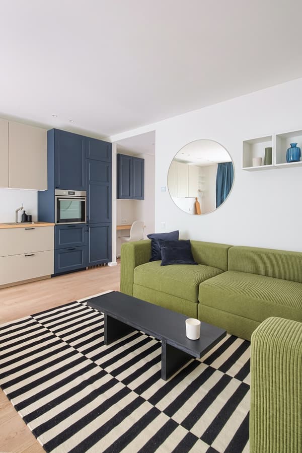 Modern and cozy open-plan living space featuring a bold green sectional sofa with navy cushions, a black coffee table, and a striped black-and-white rug. The kitchen area includes deep blue cabinetry with built-in appliances and light beige lower cabinets with wooden countertops. A round wall mirror adds depth, while open shelves display minimal decorative items. Light wood flooring and soft natural light contribute to the room’s clean, fresh look.