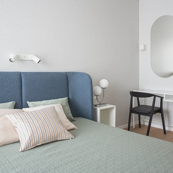 Modern and minimal bedroom featuring a blue upholstered headboard, soft green quilted bedding, and a mix of striped and solid pillows in pastel tones. A white wall-mounted reading light is positioned above the bed, while a small white side table holds a sculptural globe-style lamp. A black chair is placed at a sleek white floating desk, with a large arched mirror on the wall above, adding depth and elegance to the light-toned space.