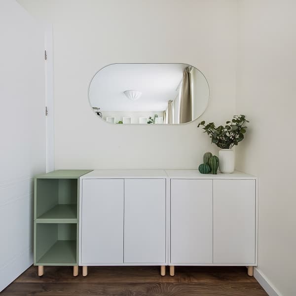 Modern and minimalistic entryway or hallway setup featuring a sleek white cabinet with natural wooden legs. On the left side, an open shelving unit in sage green adds a soft color accent. Above the cabinet, an oval wall mirror reflects a bright room with beige curtains. On the right side of the cabinet, decorative items including a white vase with green foliage and small green ceramic cacti add a fresh and natural touch. The space has a clean, Scandinavian-inspired aesthetic with soft, neutral tones and natural wood flooring.