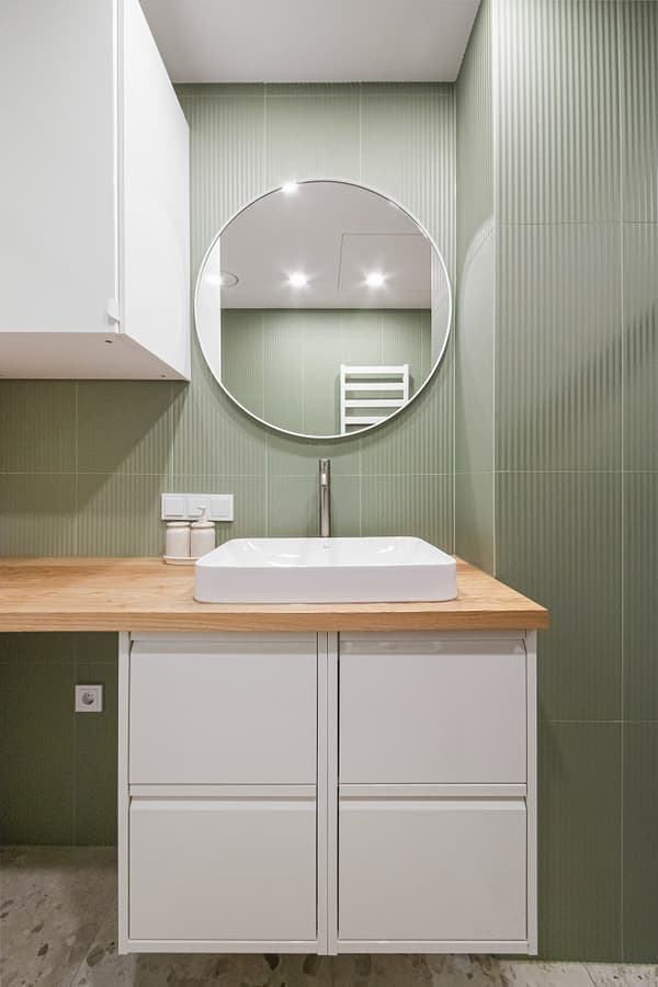 Modern bathroom vanity area with a clean and minimalist design. The space features vertical sage green tiles, a round wall mirror, and a white vessel sink mounted on a light wood countertop. Below the sink is a white floating cabinet with handle-less drawers. On the left side of the counter are matching soap dispensers, and recessed lighting above adds brightness to the serene, spa-like atmosphere.