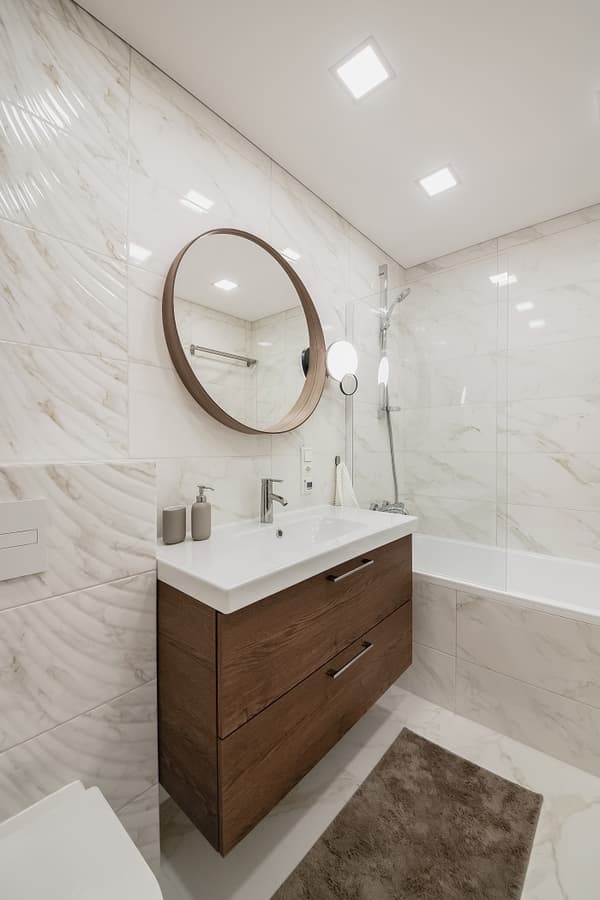 Modern bathroom with a floating wooden vanity, round mirror, wall-mounted faucet, and marble-patterned tiles.