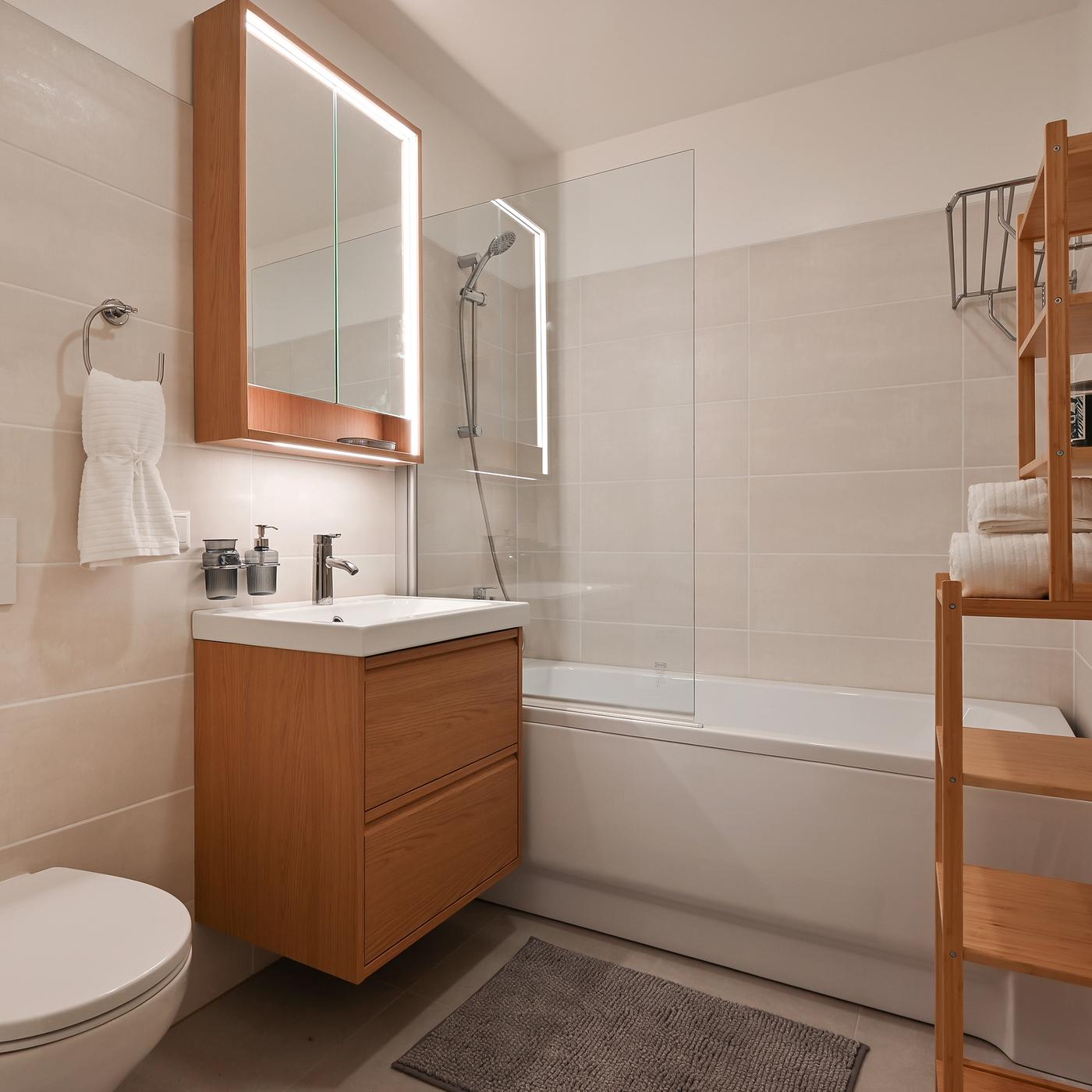 Modern bathroom with beige wall tiles, a wooden vanity unit, illuminated mirror cabinet, bathtub with glass shower screen, and open wooden shelving creating a warm and minimalist interior.