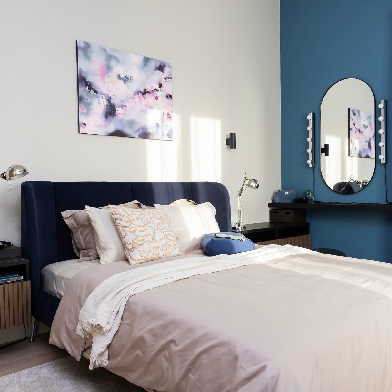 Modern bedroom with a dark blue upholstered headboard, light bedding and decorative cushions; an abstract painting hangs above the bed, and in the corner there is a blue accent wall with a round mirror and a small table.