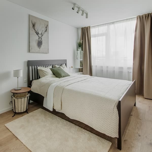 Modern bedroom with a dark wooden bed frame, cream bedding, and green accent pillows. A drum-style nightstand holds a small lamp, and a canvas art piece of a deer hangs above the bed. Natural light filters through sheer curtains paired with taupe drapes. A beige rug and wooden flooring enhance the cozy, minimalist style.