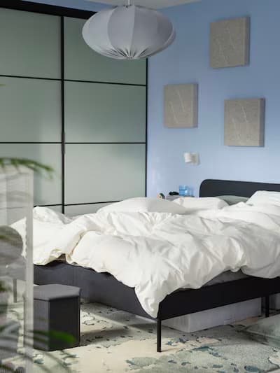 Modern bedroom with white bedding, black headboard, light blue wall, frosted glass sliding wardrobe, and white pendant light.