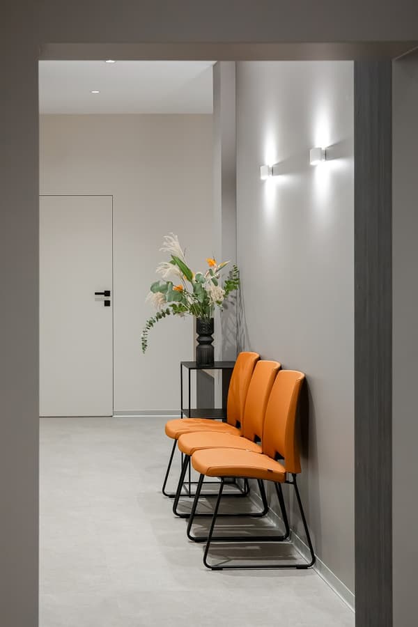 Modern dental clinic hallway with bright orange chairs, decorative floral arrangement, and minimalist wall lighting.