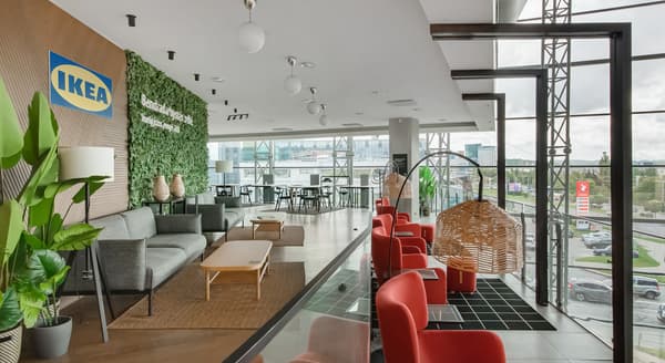 Modern IKEA lounge area with floor-to-ceiling windows, green accent wall, Scandinavian-style furniture including gray sofas, red armchairs, wicker pendant lights, and a mix of plants and natural materials in a bright showroom setting.