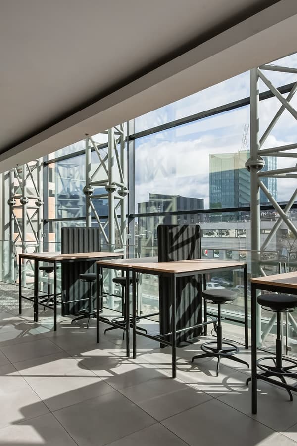 Modern IKEA workspace with high tables and bar stools, tall cushioned backrests, and floor-to-ceiling windows providing natural light and a city view through a steel-frame glass facade.