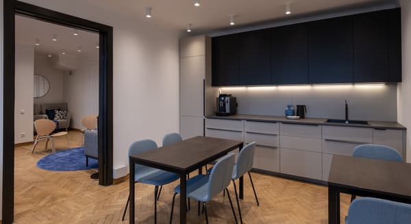 Modern kitchen and dining area featuring matte black and gray cabinetry with under-cabinet lighting. Two dark wooden tables are surrounded by light blue upholstered chairs. A coffee machine and a blue decorative vase sit on the counter. Through the open doorway, a cozy lounge area with armchairs and blue rugs is visible. The room has light-colored walls and herringbone wood flooring.