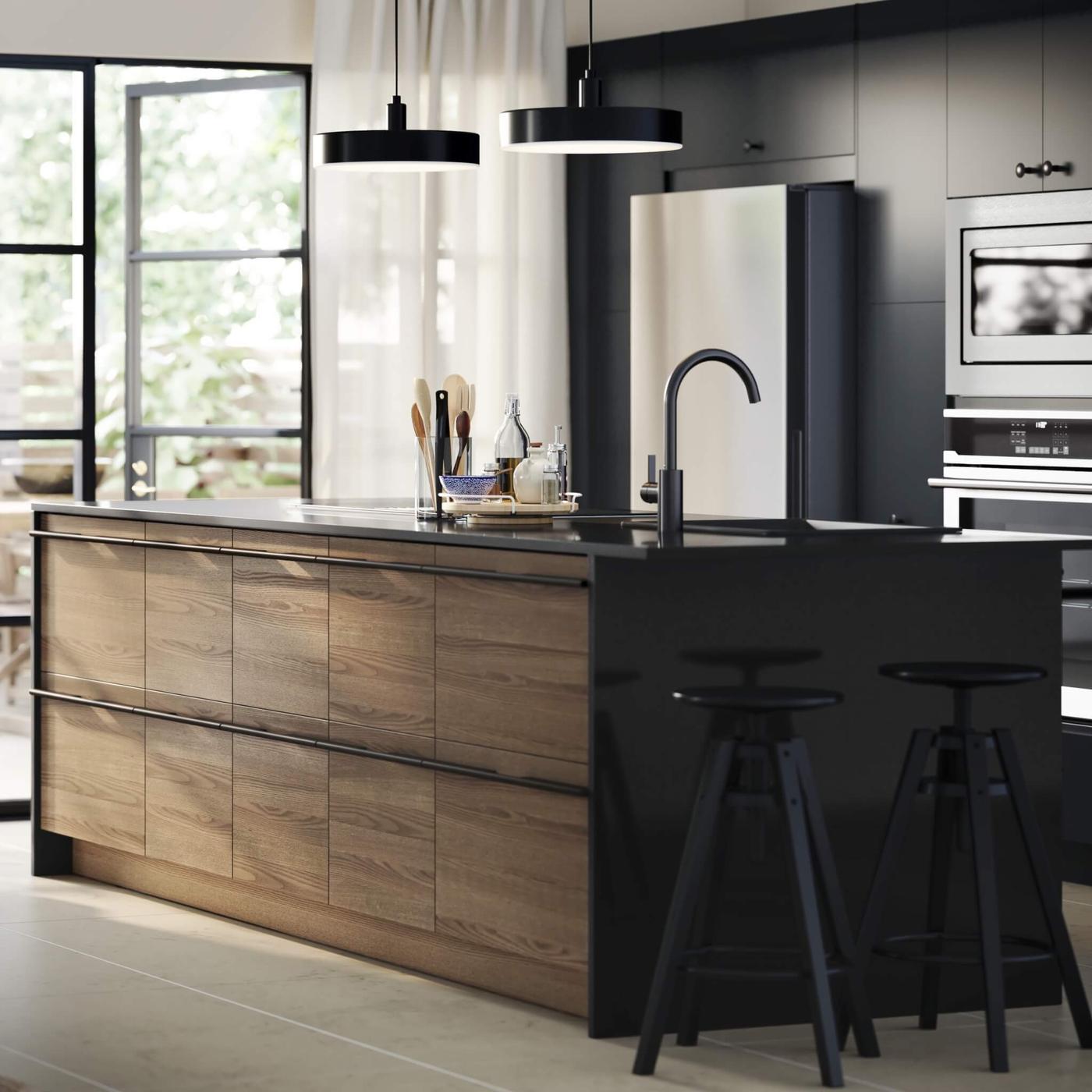 Modern kitchen with wood island, black stools, pendant lights, stainless steel appliances, and large windows with natural light.