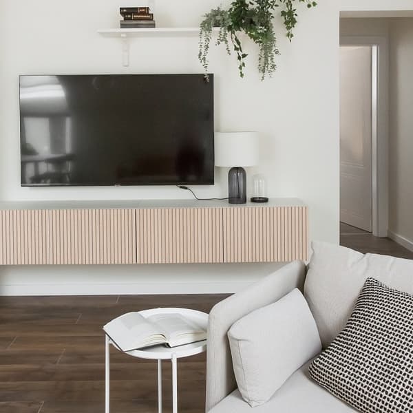 Modern living room corner with a mounted flat-screen TV above a light wood slatted floating console. A white shelf above the TV holds two stacked books and a green trailing plant. On the console, there is a table lamp with a white shade and a decorative glass dome. In the foreground, a light gray armchair with a black-and-white patterned cushion faces the TV. A small round white side table holds an open book, and the room features dark wood flooring and minimalist decor.