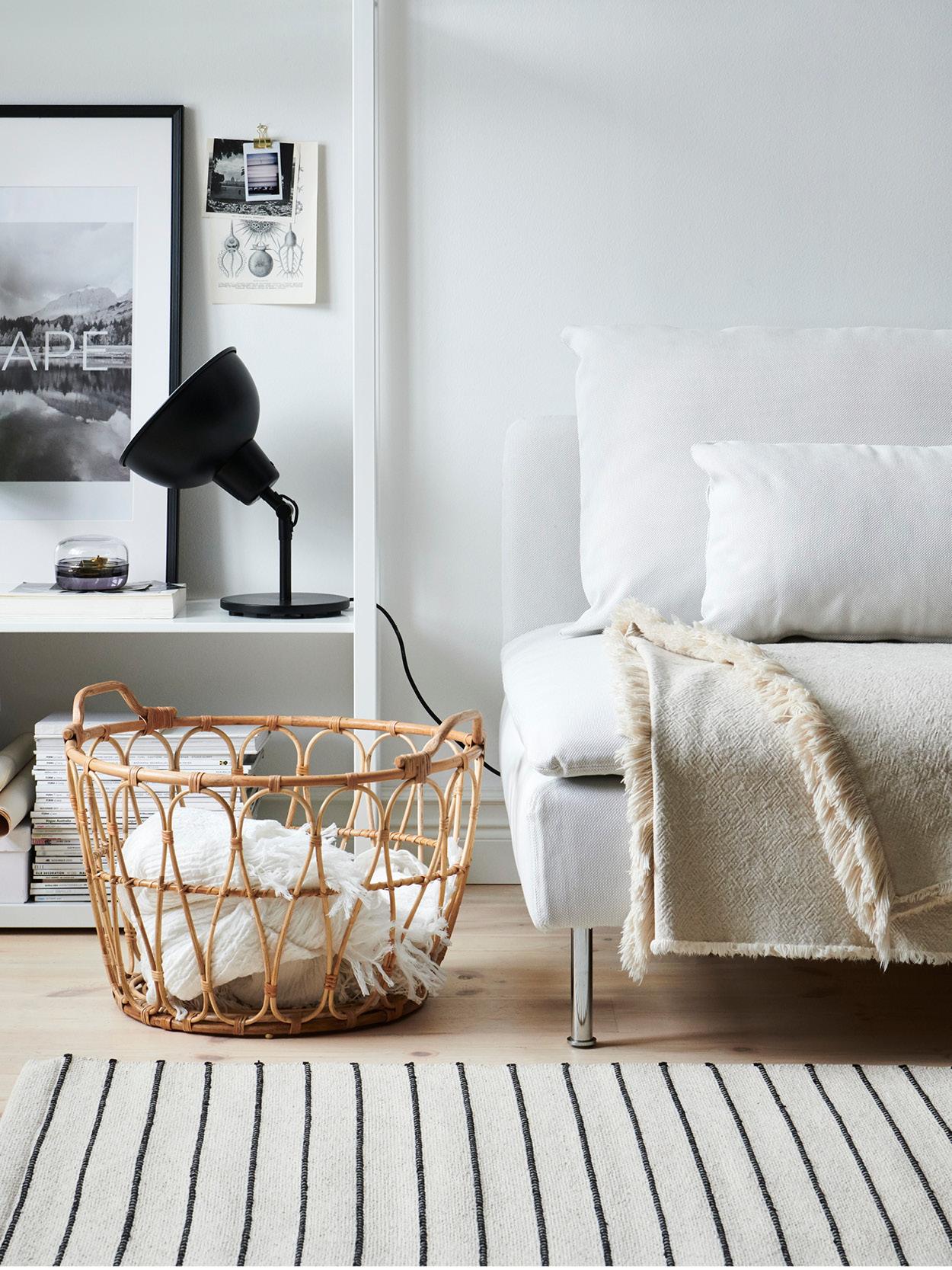 Modern living room with a light sofa, beige blanket, white cushions, white shelf with black table lamp and decorations, rattan basket with blankets, wall art and a light carpet with black stripes.