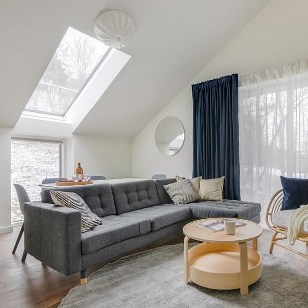 Modern living room with a sloped ceiling and large skylight letting in natural light. A dark gray sectional sofa with assorted pillows sits on a soft gray rug. In front of the sofa is a light wood round coffee table with a candle and magazine. Blue blackout curtains and sheer white drapes cover a large window, while a round mirror decorates the white wall. In the corner, a rattan chair with a dark blue pillow and throw adds a cozy touch. The space has a clean, Scandinavian-inspired aesthetic.