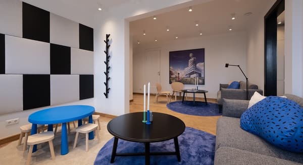 Modern lounge and play area featuring a cozy seating space with gray sofas, blue accent pillows, and round black coffee tables placed on soft blue rugs. To the left, a child-sized blue play table with wooden stools stands near a wall decorated with large black and gray acoustic panels. A black coat rack shaped like branches is mounted on the wall. In the background, a wall art piece of a futuristic building scene adds a creative touch. The room has parquet wood flooring and is lit with multiple ceiling spotlights.