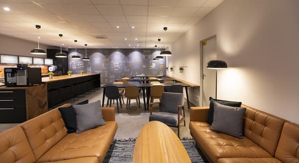 Modern office lounge with brown leather sofas, grey armchairs, wooden coffee table, and open kitchen in the background with pendant lights.