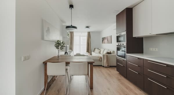 Modern open-concept kitchen and living room with a warm, minimalist design. The kitchen features sleek dark wood lower cabinets and integrated appliances, contrasted by white upper cabinets. A wooden dining table with white chairs separates the kitchen from the living area. Soft brown curtains, a neutral-toned sectional sofa, and soft lighting create a cozy ambiance.
