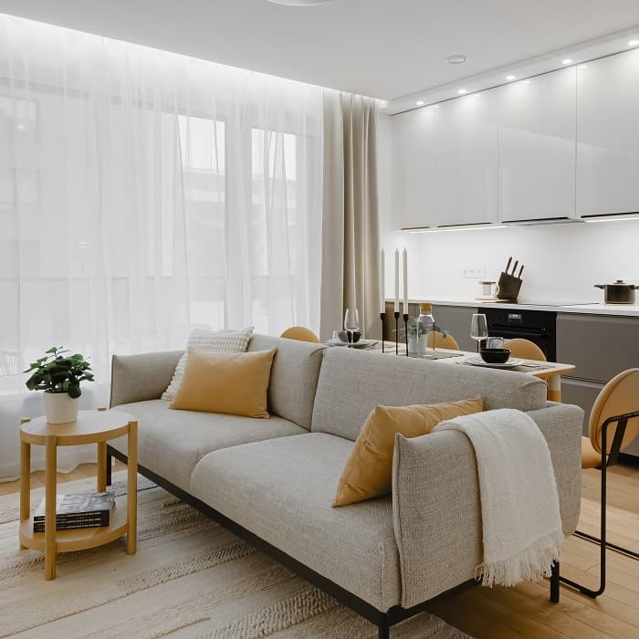 Modern open-concept living room and kitchen with a light gray sofa featuring mustard yellow throw pillows, a white throw blanket, and a round wooden side table with a potted plant and magazines. Sheer white curtains allow natural light to filter in, and the kitchen in the background has sleek white cabinets, under-cabinet lighting, and a dining table set for two with tall candles and black dinnerware.