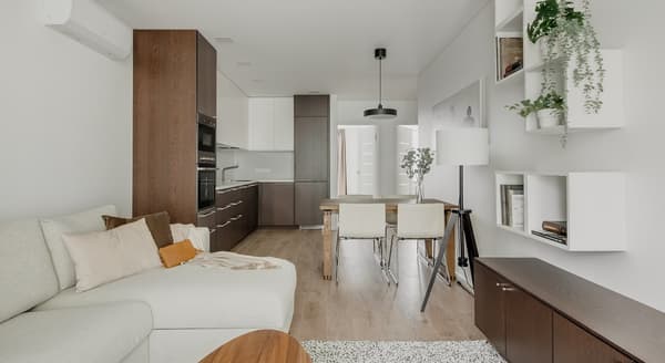 Modern open-concept living room and kitchen with a minimalist Scandinavian design. The space features a light beige sectional sofa with neutral pillows, a wooden dining table with four white chairs, sleek dark wood and white kitchen cabinetry, and floating white shelves with green plants and books. The room is bright, with light wood flooring and natural light coming from the hallway.