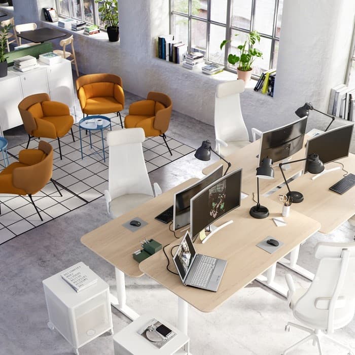 Modern open-plan office with light wood desks, white ergonomic chairs, and multiple computer monitors and task lamps. A laptop and various office supplies are placed on the desks. In the background, there is a cozy lounge area with mustard-colored armchairs and small round tables on a checkered rug. Large windows allow natural light to fill the space, with books and potted plants on the windowsills.