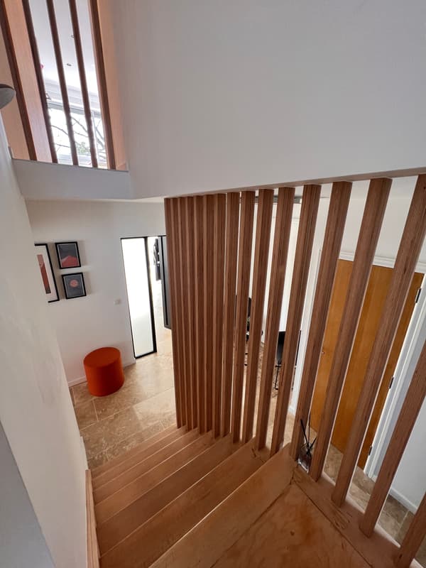 Moderne houten trap met verticale spijlen die uitkomt in een lichte hal met witte muren en een rood krukje.