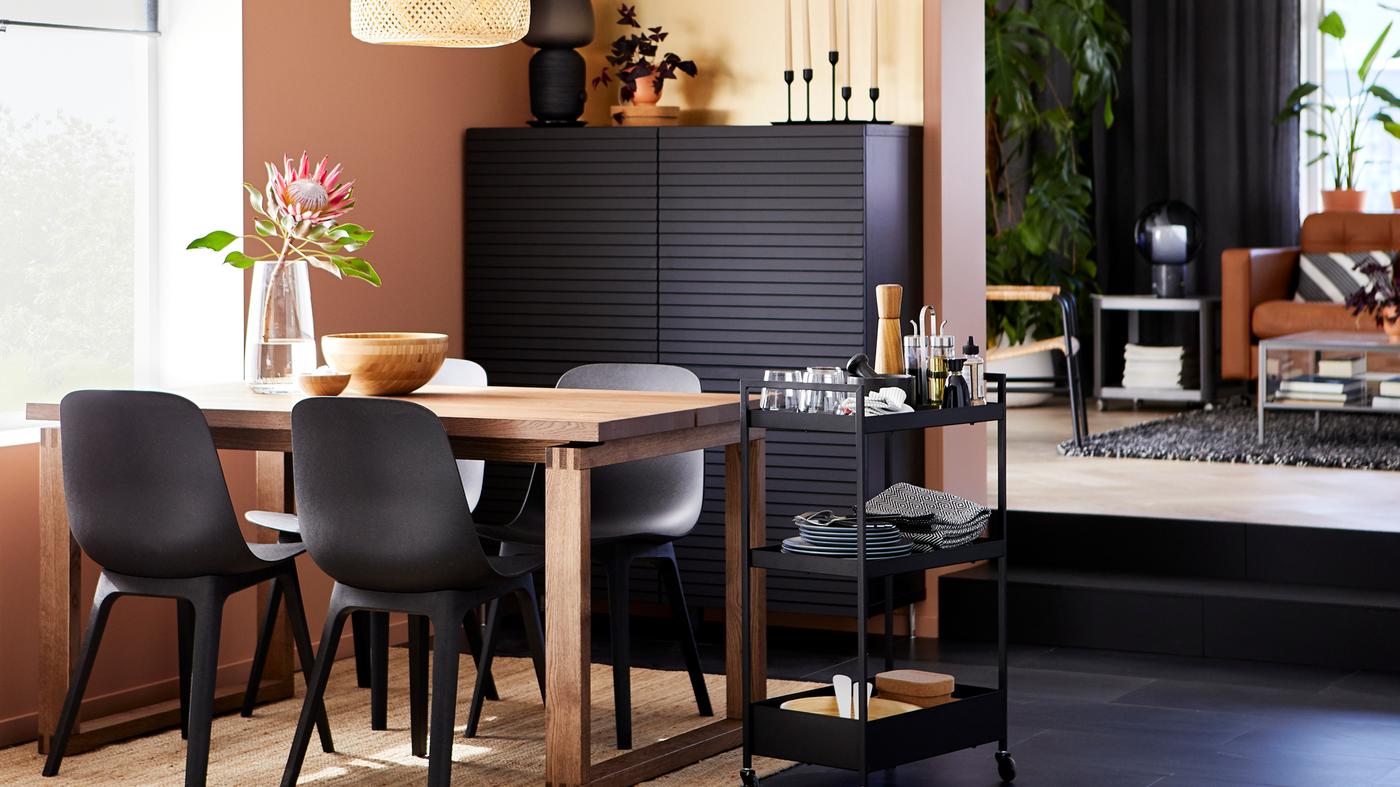 MÖRBYLÅNGA oak veneer table with black ODGER chairs by a window, with a black cabinet and a trolley holding tableware.