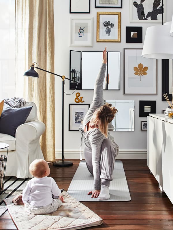 Mother doing yoga on a mat in a living room with a baby sitting on a mat watching her with sofa, sideboard and pictures.