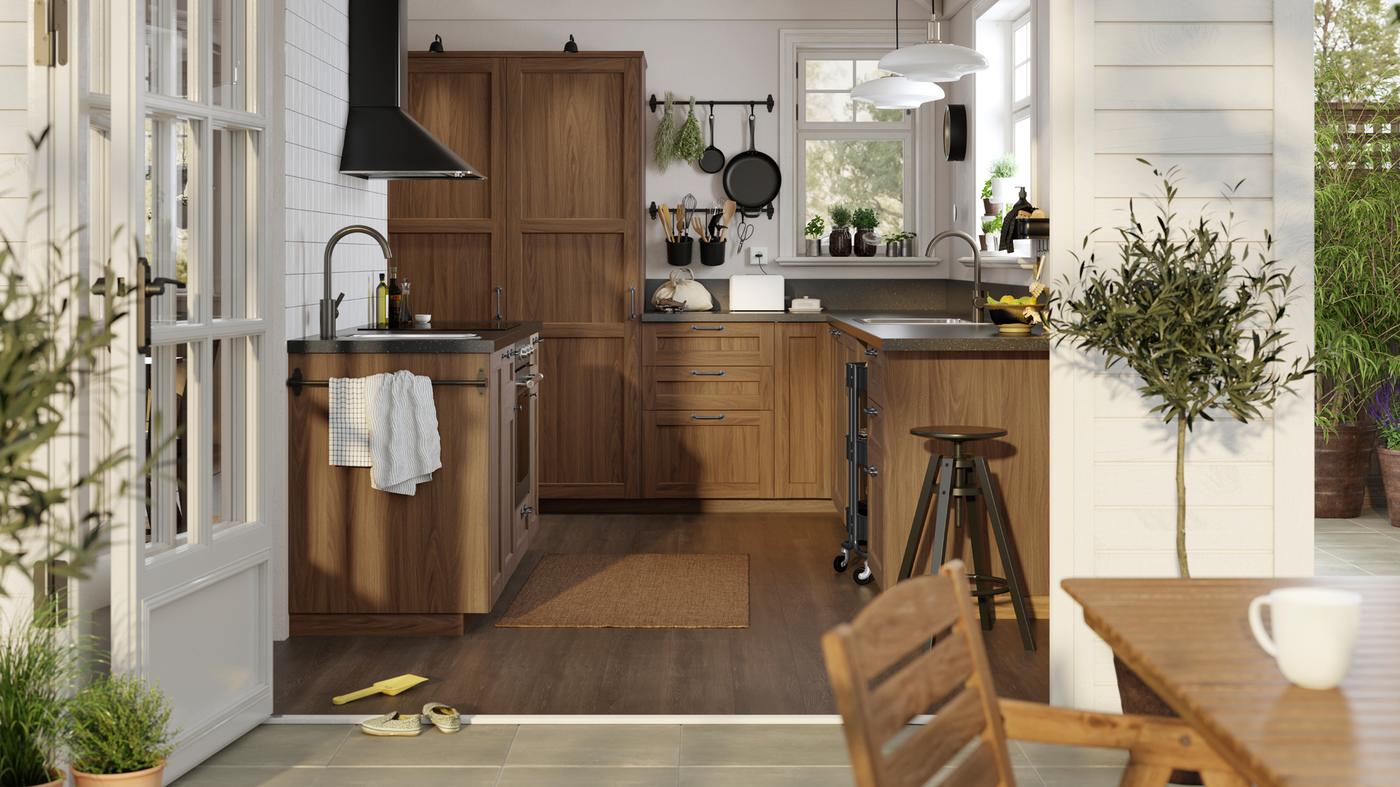 مطبخ مشمس أبيض اللون بتصميم كلاسيكي مع واجهات أدراج بني شكل خشب الجوز ENKÖPING وأسطح عمل سوداء بتأثير معدني.