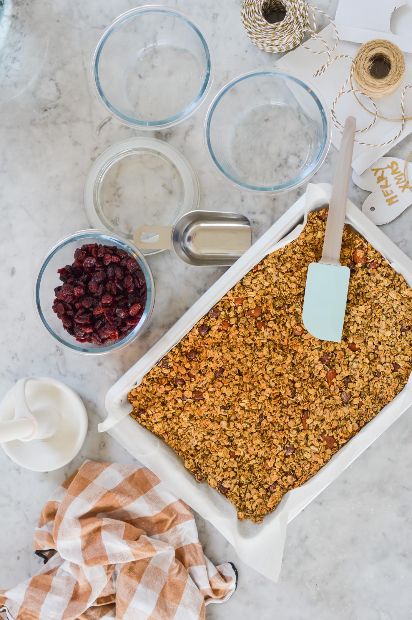 Muesli in una pirofila con una spatola blu su un piano di lavoro in marmo. Circondato da ciotole con mirtilli rossi secchi, barattoli di vetro e un panno a righe, che evocano un'accogliente scena di cucina.