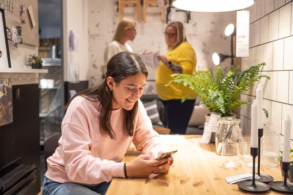 Mujer sentada en una mesa con el celular en la mano, atras dos mujeres hablando dentro de un espacio en la tienda IKEA.