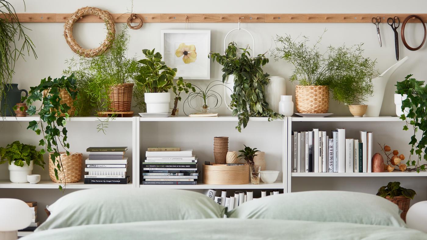 Multiple potted plants, real and artificial, stand amid books and decoration on shelves by a bed with BERGPALM bed linen.