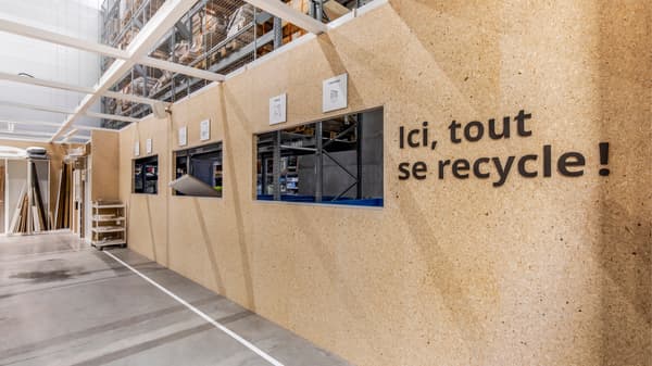 Mur en panneaux recyclés avec guichets et texte « Ici, tout se recycle », dans un espace logistique lumineux.