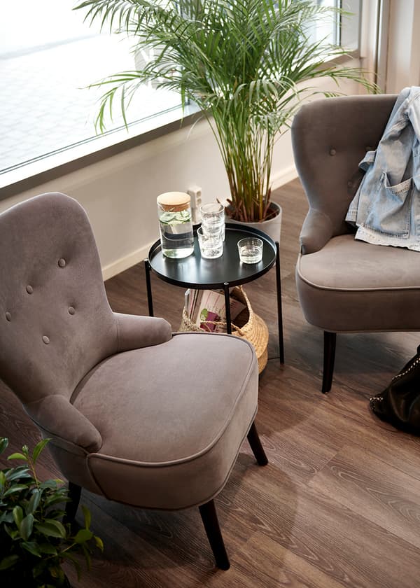 Mysig hörna med två grå stolar, ett sidobord med vattenkanna, glas och ljus . En växt står bakom och en jacka hängd över ena stolen.

