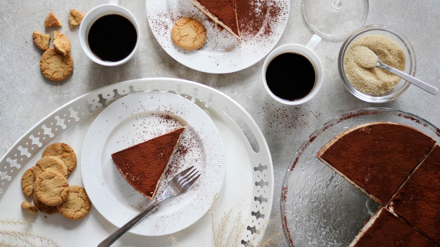 Na servírovacím stojanu ARV BRÖLLOP je cheescake posypaný kakaem, vedle leží dva dezertní talíře OFANTLIGT s jednotlivými porcemi, dva hrnky s kávou a cukřenka s třtinovám cukrem.