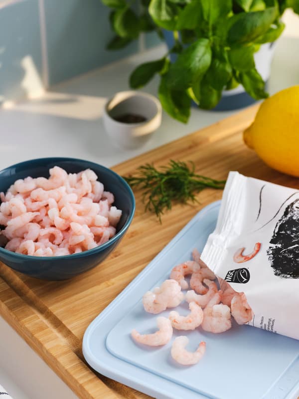 Načatý sáček krevet SJÖRAPPORT leží na bleděmodrém prkénku IKEA 365+, několik krevet je vysypáno, vedle stojí tmavě tyrkysová miska FÄRGKLAR plná krevet, v pozadí je celý citron a svazek bazalky.