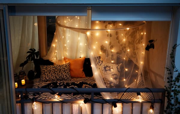 Nächtlicher Blick von draussen auf einen geschlossenen Balkon, dekoriert wie ein Schlafzimmer, u. a. mit SVARTRÅ Lichterketten (12), LED schwarz/für draussen.
